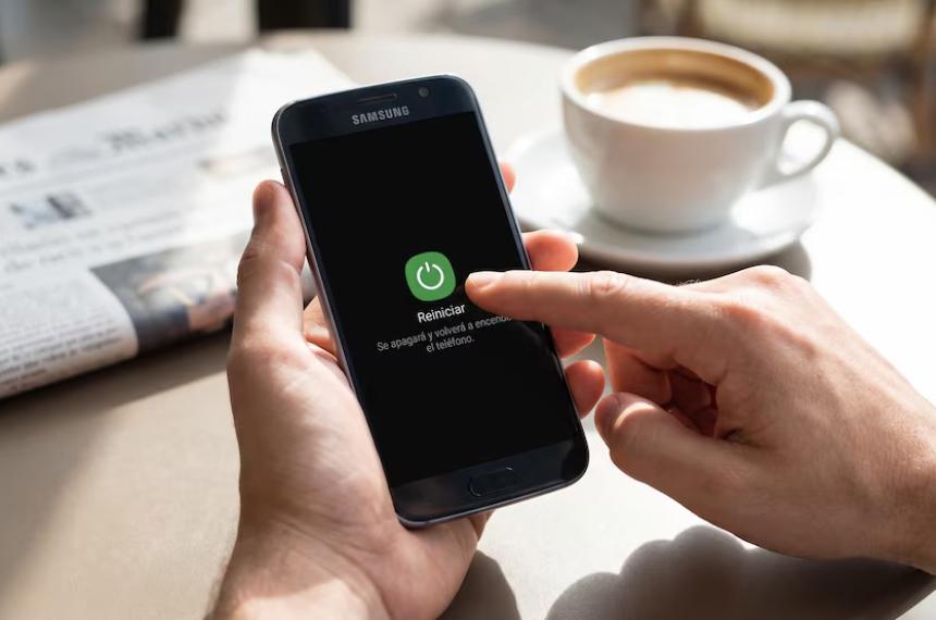Una persona reinicia su teléfono Samsung con la opción Reiniciar visible en pantalla mientras un periódico y una taza de café se encuentran desenfocados en segundo plano