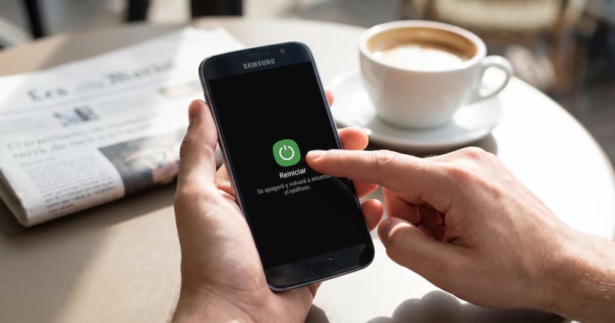 Una persona reinicia su teléfono Samsung con la opción Reiniciar visible en pantalla mientras un periódico y una taza de café se encuentran desenfocados en segundo plano